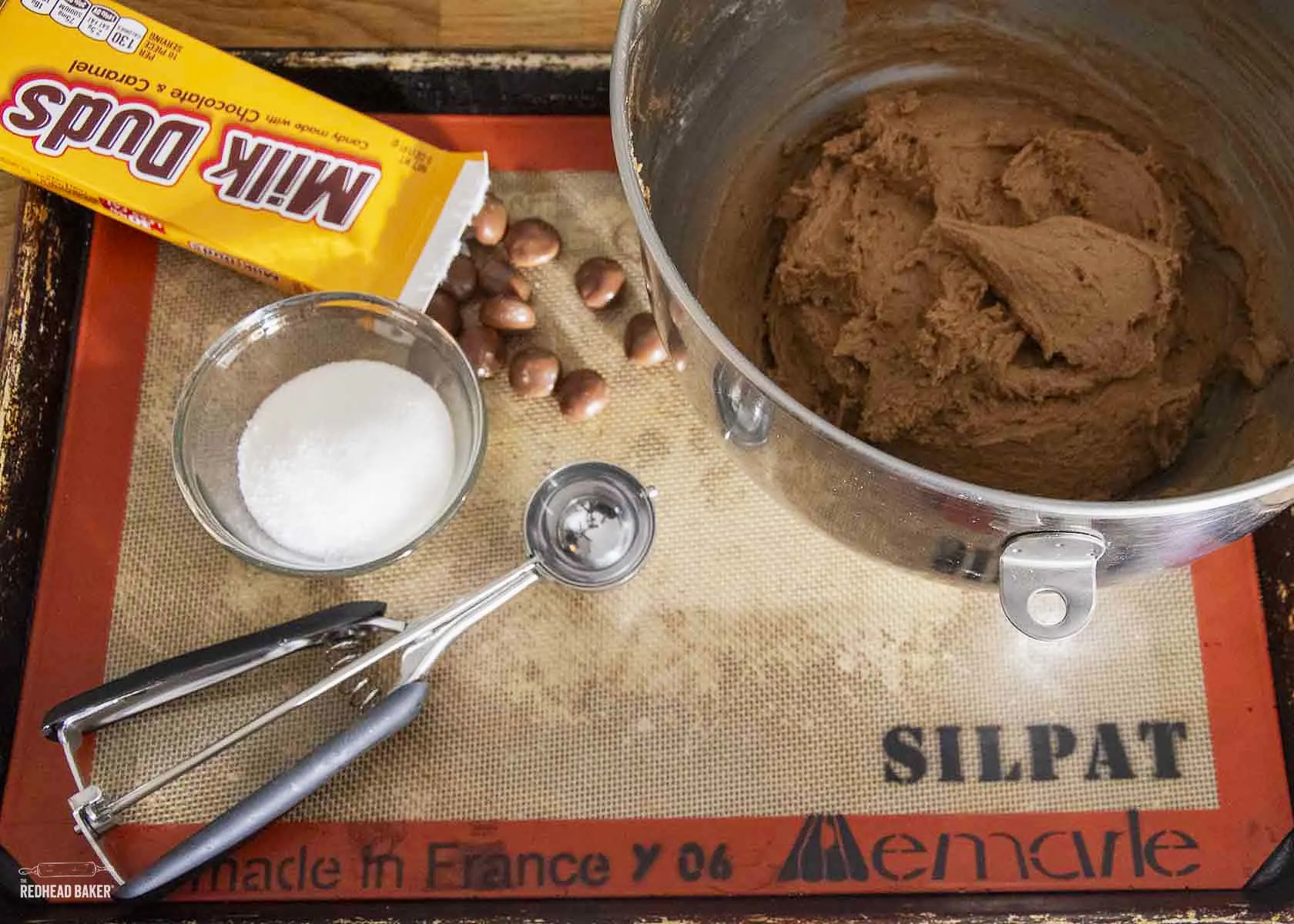 The bowl of cookie dough, a cookie scoop, a bowl of sugar and a box of Milk Duds on a silicone mat-lined baking sheet.