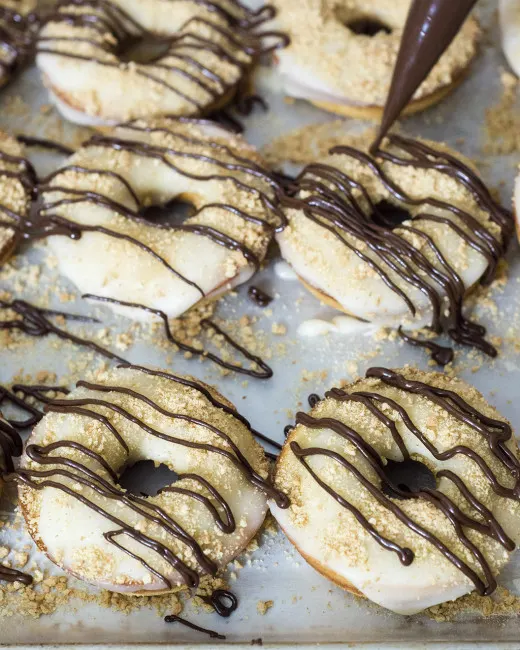 S'mores in donut form &mdash; are they for breakfast or dessert? Baked vanilla donuts are iced in marshmallow glazed, then dusted in graham cracker crumbs and a chocolate drizzle. TheRedheadBaker.com #WhatsBaking