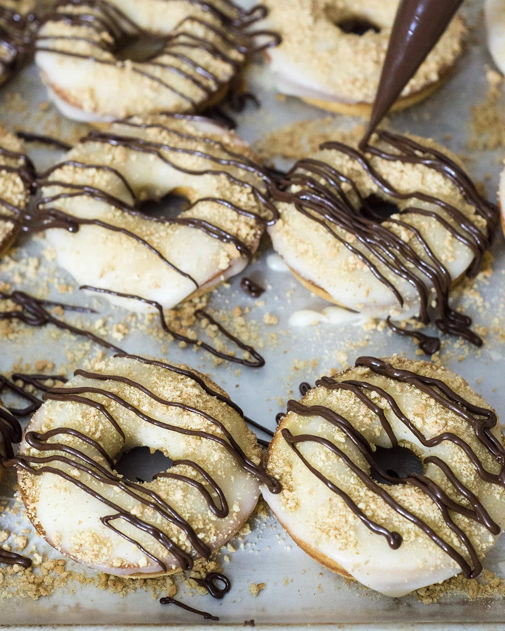 S'mores in donut form — are they for breakfast or dessert? Baked vanilla donuts are iced in marshmallow glazed, then dusted in graham cracker crumbs and a chocolate drizzle. TheRedheadBaker.com #WhatsBaking