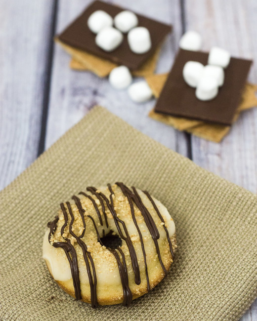 S'mores in donut form — are they for breakfast or dessert? Baked vanilla donuts are iced in marshmallow glazed, then dusted in graham cracker crumbs and a chocolate drizzle. TheRedheadBaker.com #WhatsBaking