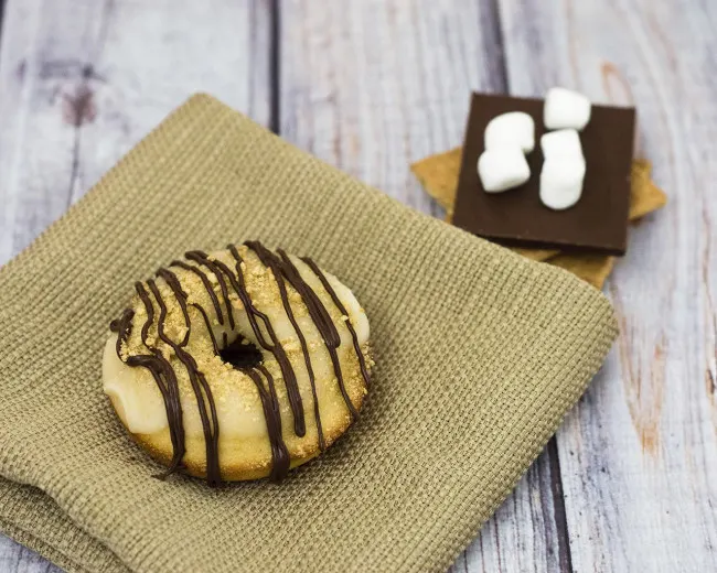 S'mores in donut form &mdash; are they for breakfast or dessert? Baked vanilla donuts are iced in marshmallow glazed, then dusted in graham cracker crumbs and a chocolate drizzle. TheRedheadBaker.com #WhatsBaking