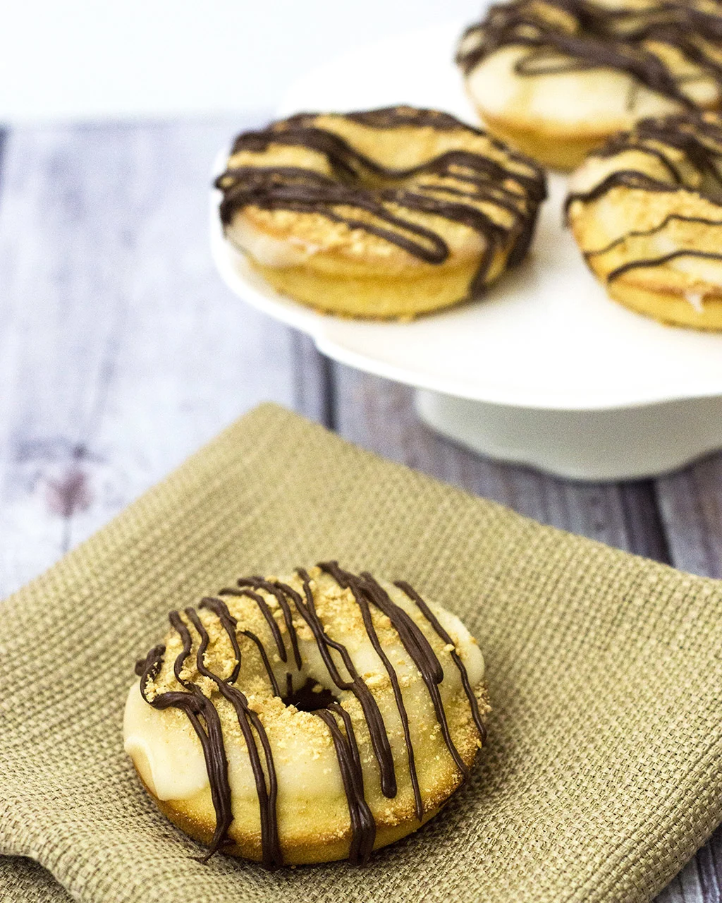 S'mores in donut form — are they for breakfast or dessert? Baked vanilla donuts are iced in marshmallow glazed, then dusted in graham cracker crumbs and a chocolate drizzle. TheRedheadBaker.com #WhatsBaking