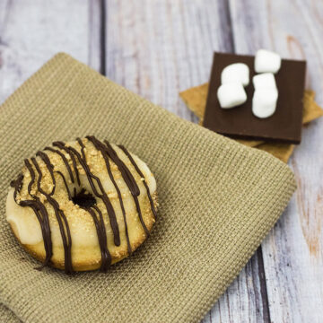 S'mores in donut form — are they for breakfast or dessert? Baked vanilla donuts are iced in marshmallow glazed, then dusted in graham cracker crumbs and a chocolate drizzle. TheRedheadBaker.com #WhatsBaking