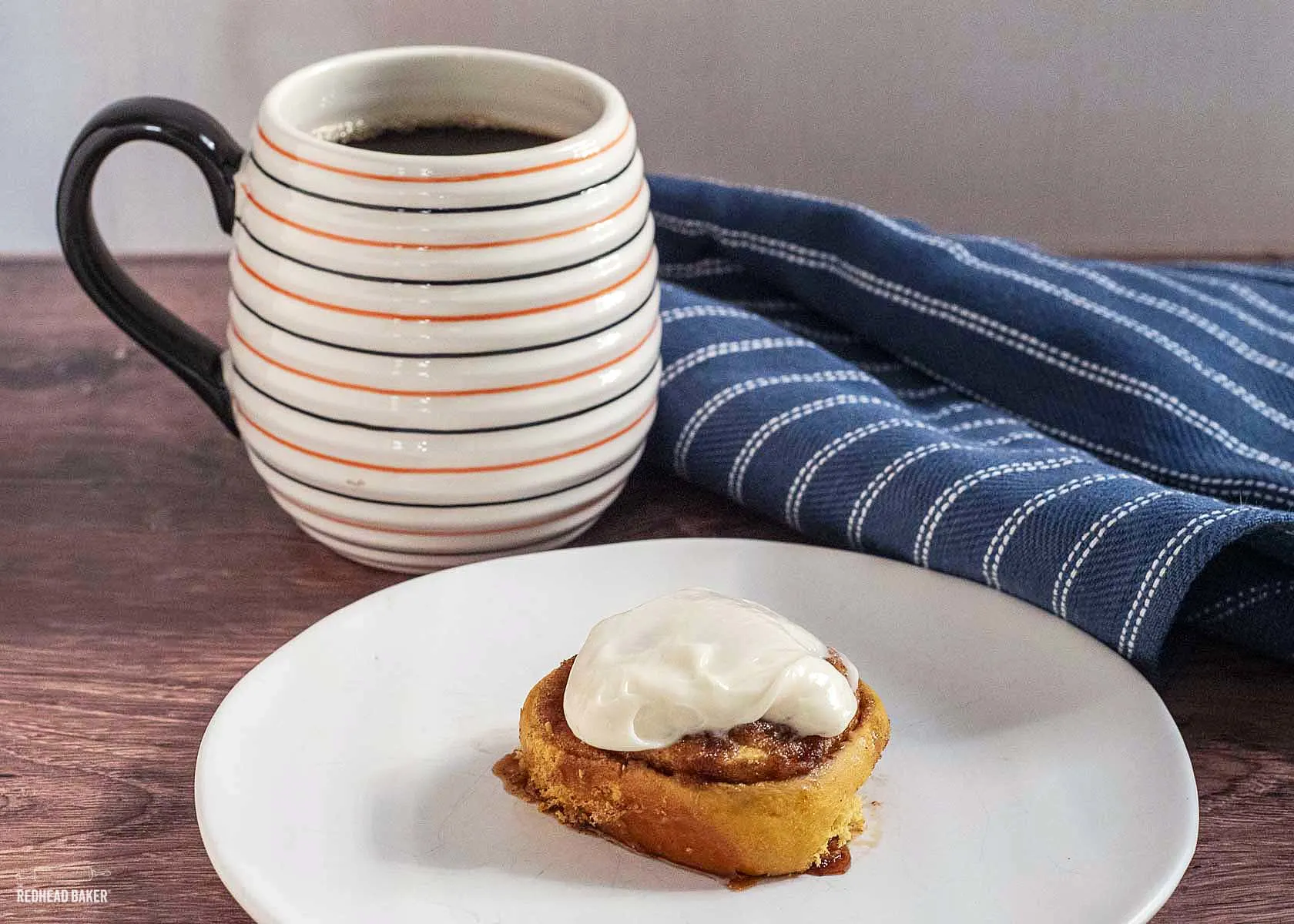 A pumpkin cinnamon roll in front of a cup of coffee. 