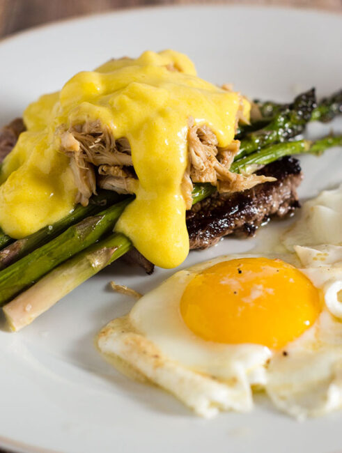 The classic brunch staple of steak and eggs gets fancy! The steak is served Oscar-style with asparagus, crab meat and Hollandaise sauce, with two sunny side-up eggs. #BrunchWeek