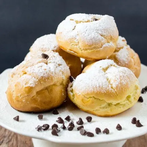A plate of powdered-sugar-dusted cannoli cream puffs with mini chocolate chips scattered around.