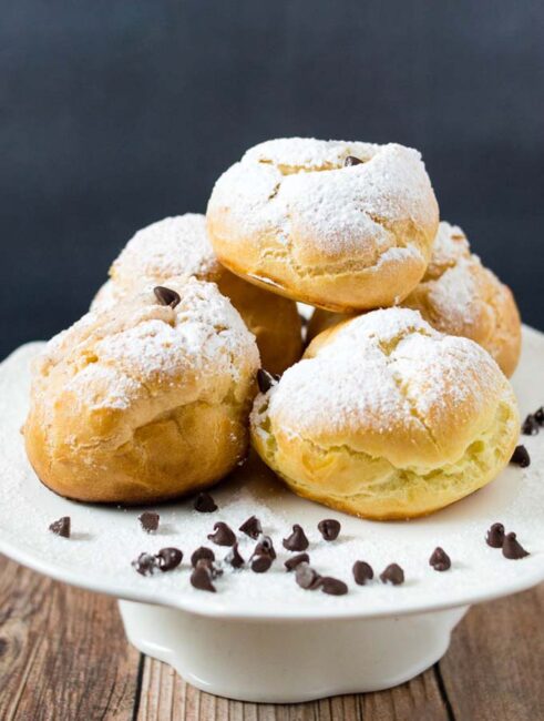 A plate of powdered-sugar-dusted cannoli cream puffs with mini chocolate chips scattered around.