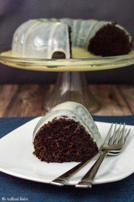A slice of fudgy chocolate bundt cake sitting in front of a cake stand holding the rest of the bundt cake.