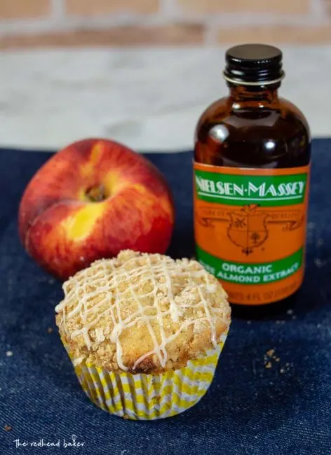 A bakery-style peach crumb muffin, a fresh peach, and a bottle of Nielsen-Massey Organic Almond Extract.
