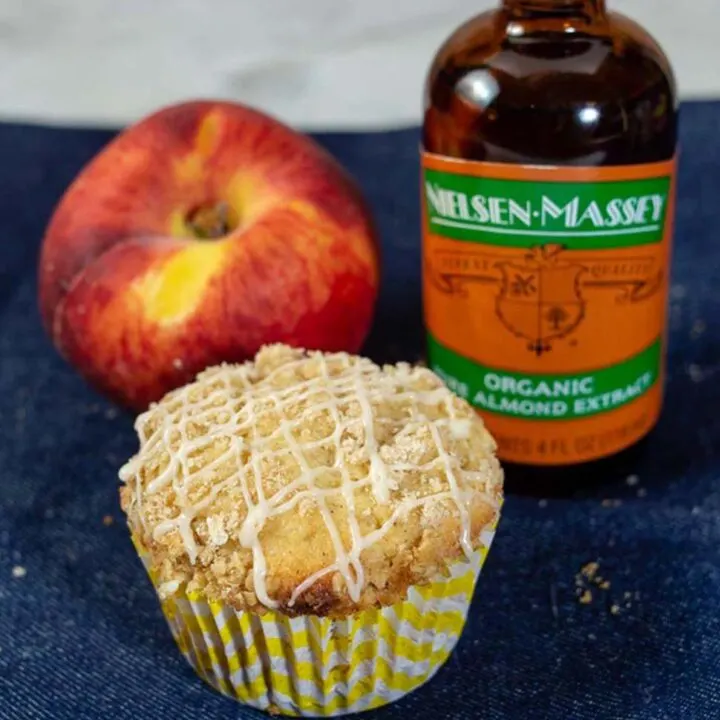 A bakery-style peach crumb muffin, a fresh peach, and a bottle of Nielsen-Massey Organic Almond Extract
