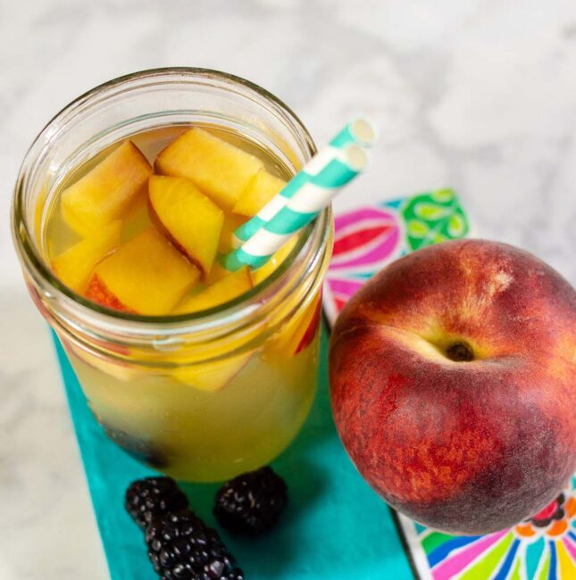 An overhead shot of a glass of sparkling blackberry peach sangria, a fresh peach and three blackberries.