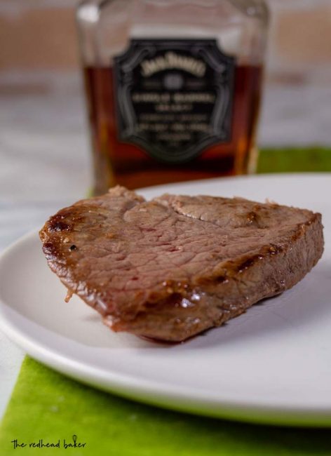 A piece of pan-seared steak in front of a bottle of whiskey.