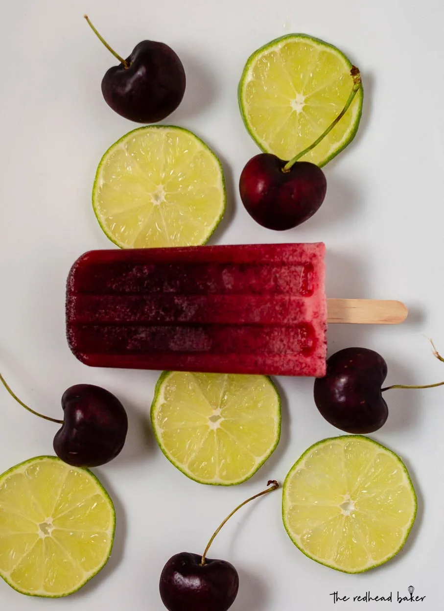 A cherry limeade pop on a white serving dish with lime slices and cherries.