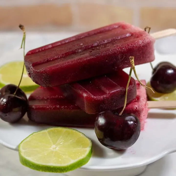 Three cherry limeade pops stacked on top of each other on a mini dessert stand with lime slices and cherries.