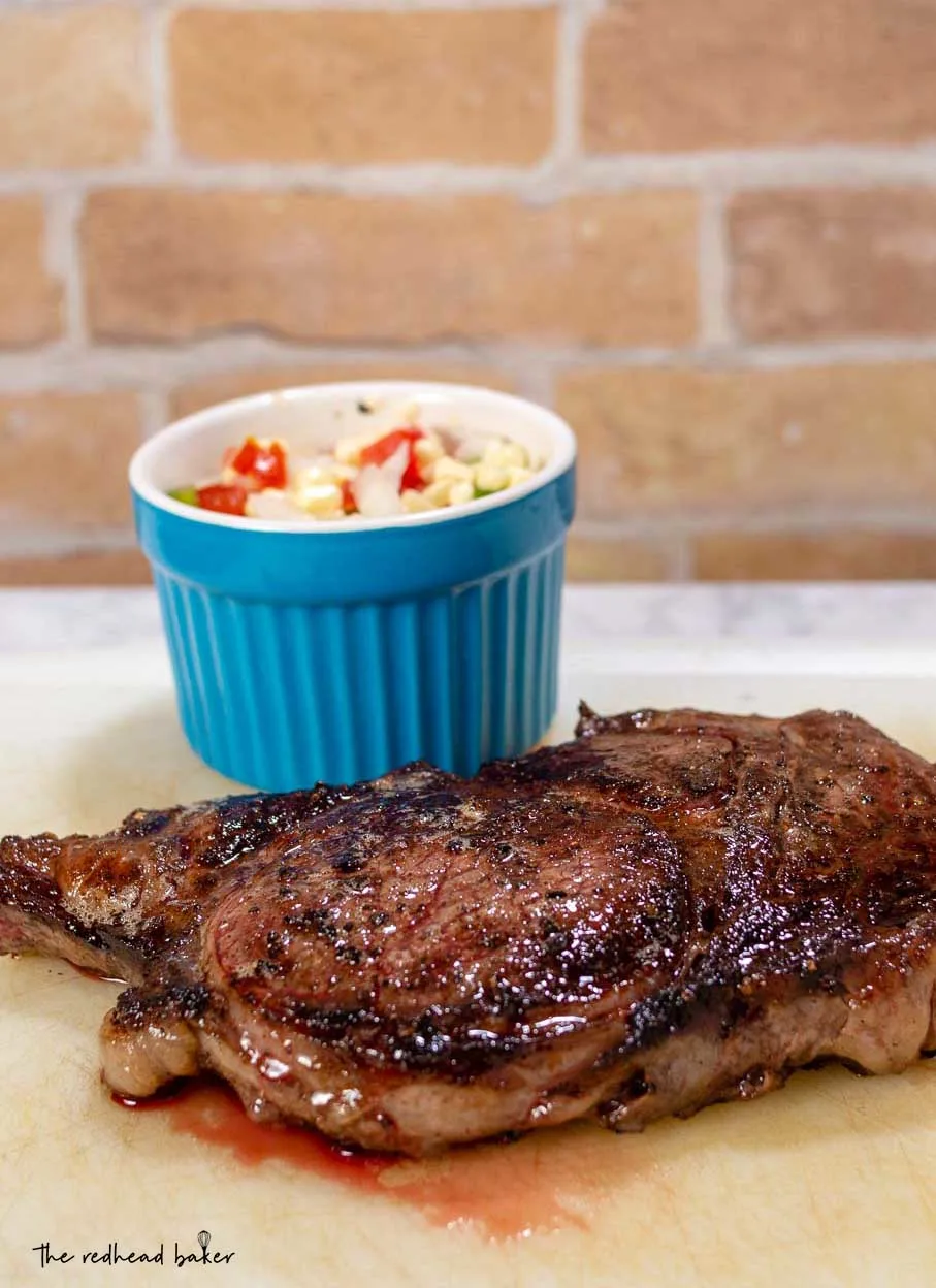 A ribeye steak resting on a cutting board with a ramekin of corn salsa behind it.