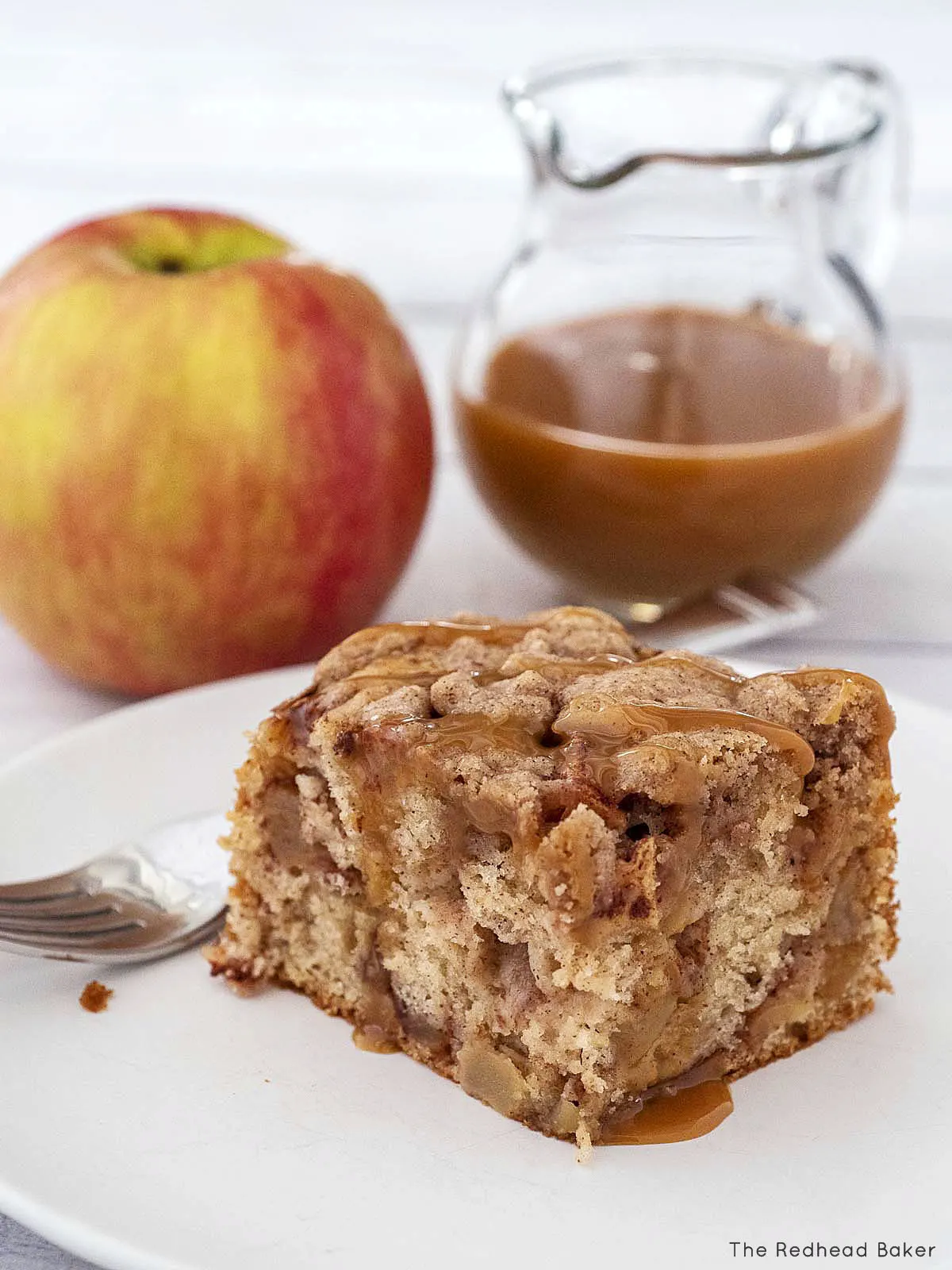 A slice of caramel apple crumb cake on a white plate in front of a jug of caramel sauce and a whole apple.