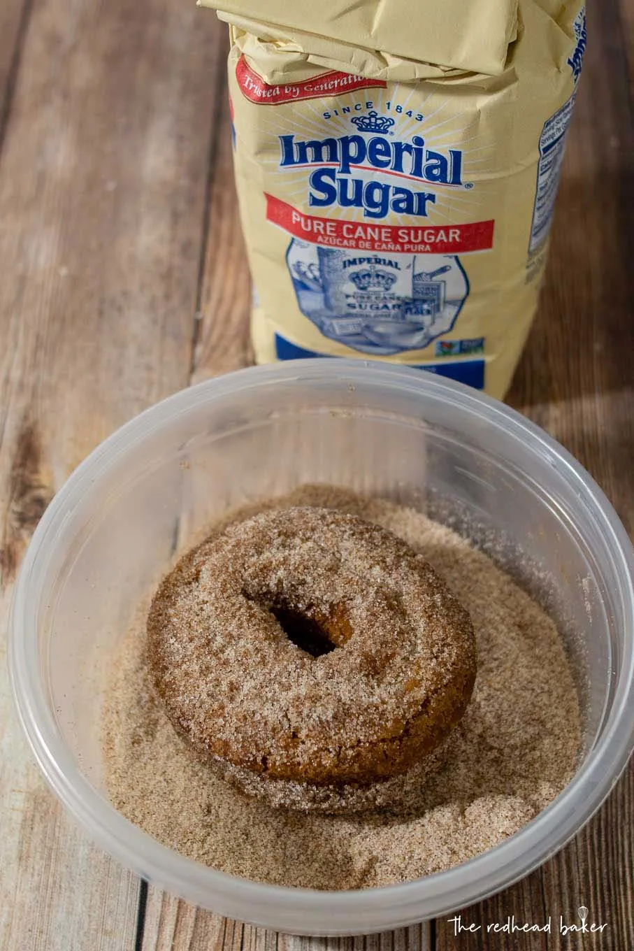 An apple cider doughnut in a dish of cinnamon sugar in front of a bag of Imperial Sugar.