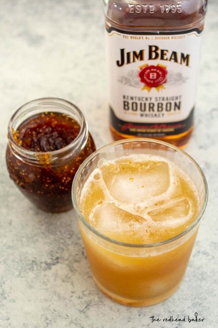 A glass of honey-fig whiskey sour in front of a jar of fig spread and a bottle of whiskey