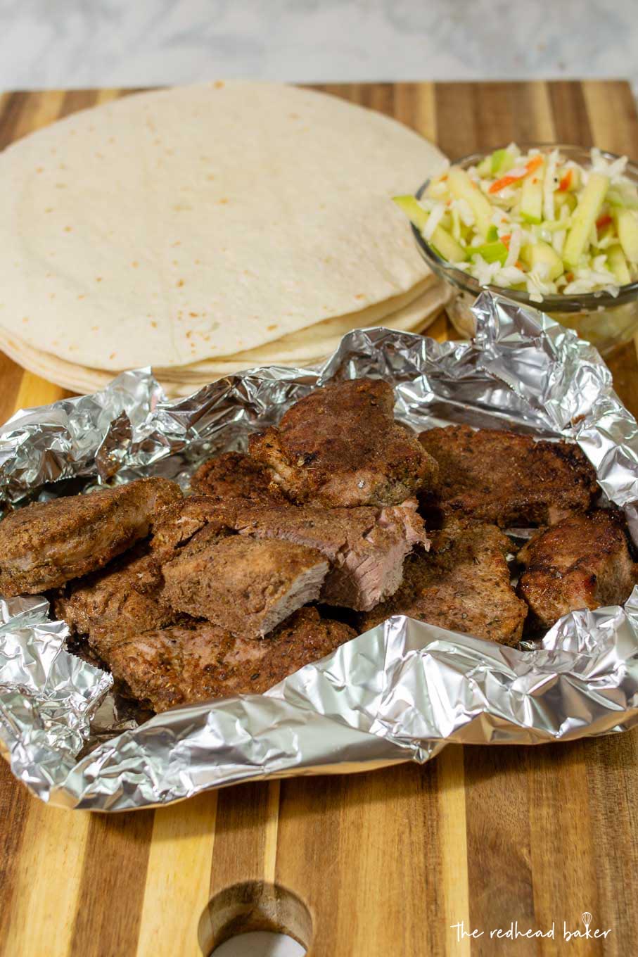 A stack of tortillas, a bowl of apple slaw, and a tinfoil pouch of pork taco meat