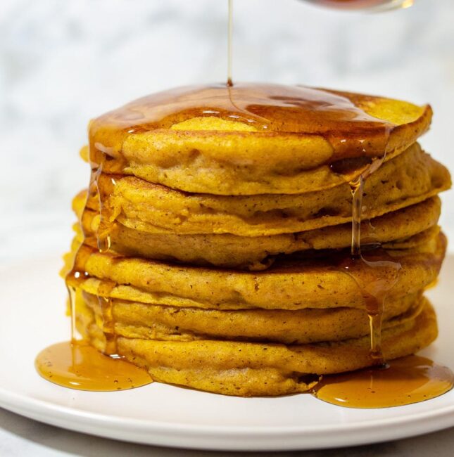 Maple syrup being poured onto a stack of pumpkin buttermilk pancakes
