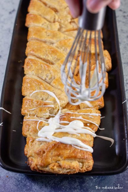 A whisk drizzling icing on an uncut pumpkin cheesecake danish braid.