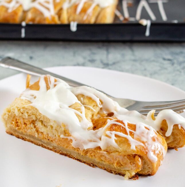 A slice of pumpkin cheesecake danish braid on a white plate in front of a black serving tray with the remaining braid.