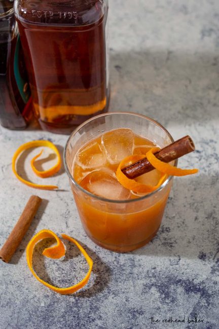 A glass of pumpkin old-fashioned in front of a bourbon bottle, orange peel twists and a cinnamon stick.