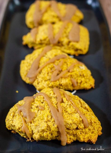 Four caramel-glazed spiced pumpkin scones on a black tray.