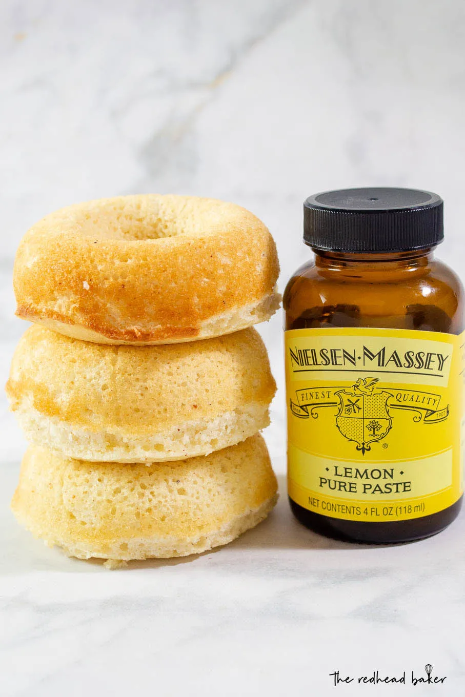 A stack of three baked lemon donuts next to a jar of Nielsen-Massey lemon paste