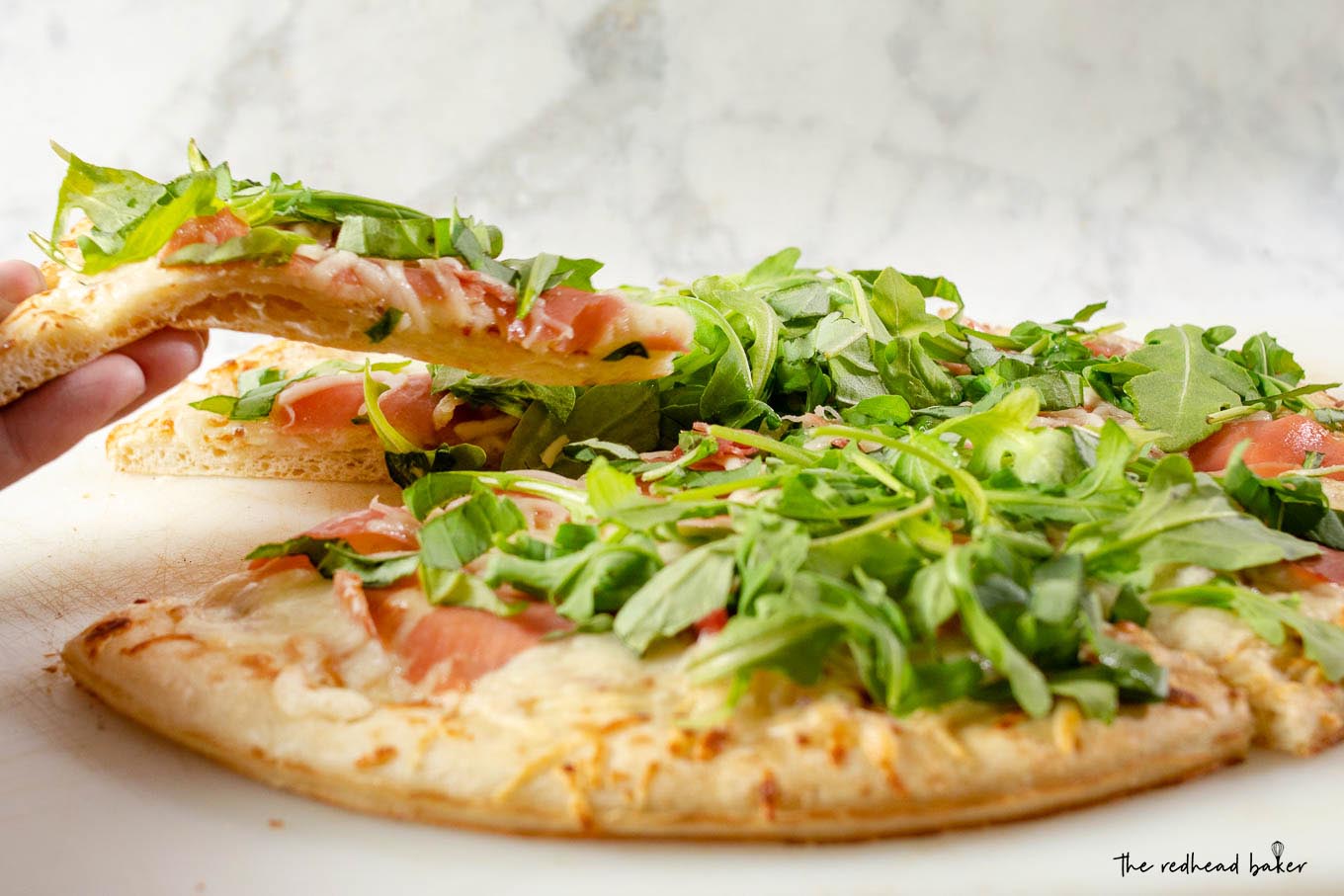 A slice of white pizza with prosciutto and arugula being lifted from the rest of the pizza