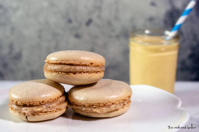 Three eggnog macarons on a cake plate in front of a mason jar of eggnog