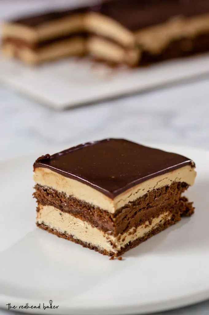 A slice of chocolate opera cake with the rest of the cake in the background
