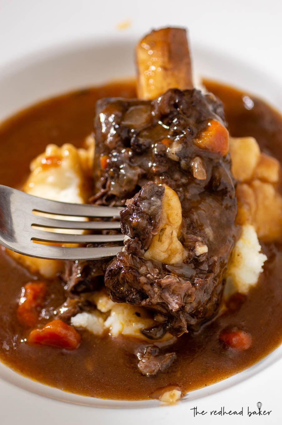 A fork with a piece of short rib on it, with the bowl of short ribs and gravy in the background.