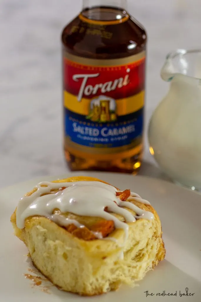 A salted caramel peach sweet roll on a white plate in front of a bottle of Torani Salted Caramel Syrup and a pitcher of glaze