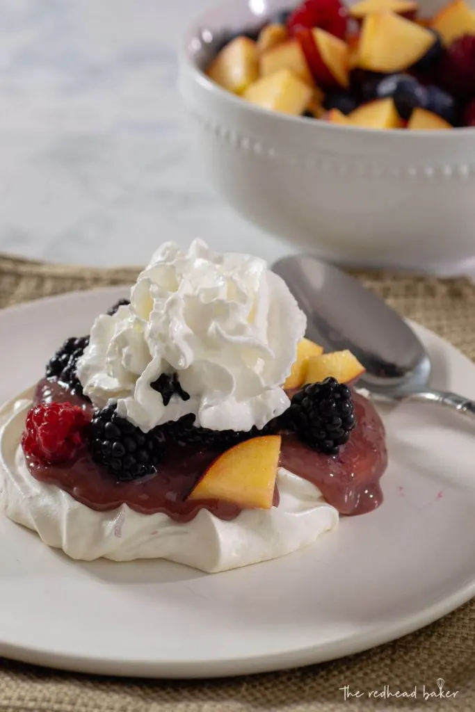A cherry-lime pavlova topped with whipped cream in front of a bowl of fresh fruit