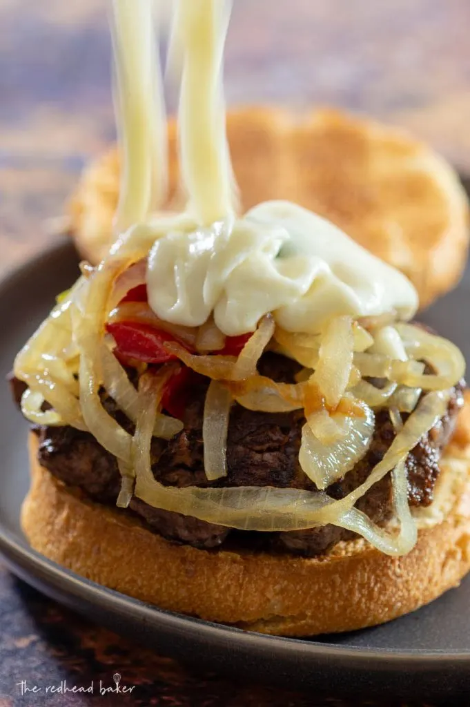 Cheese sauce being spooned onto a Philly burger topped with fried onions and peppers