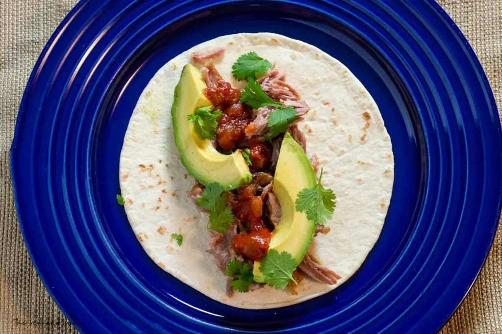 A plate with a flour tortilla, pork carnitas, avocado and cilantro