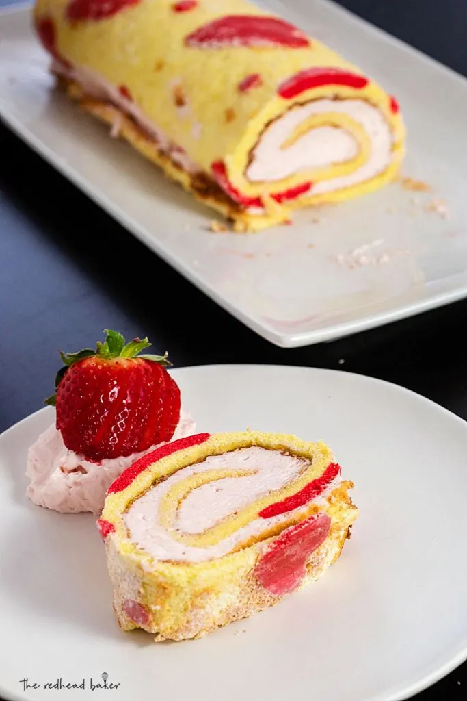 A slice of jelly roll cake on its side on a plate, with the rest of the cake roll in the background