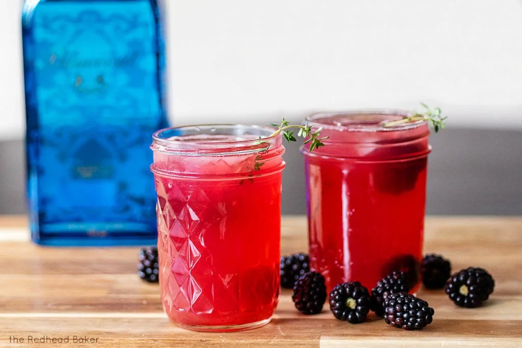 Two glasses of blackberry thyme collins on a wooden board with a bottle of gin