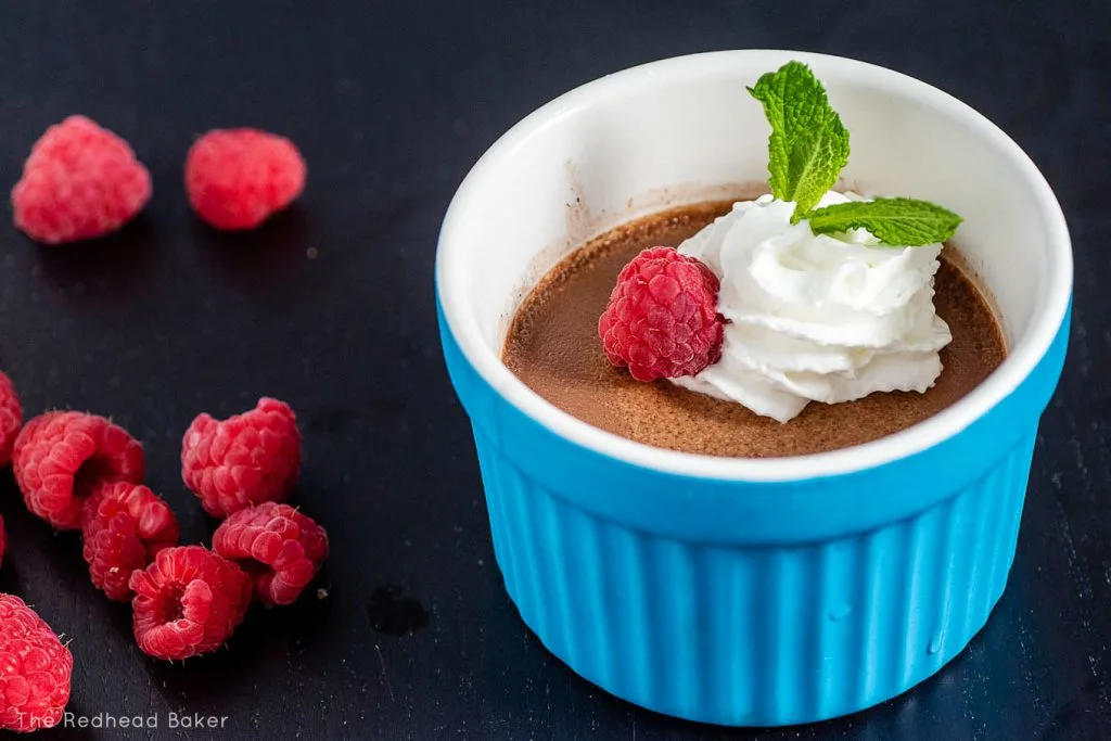 A ramekin of chocolate custard garnished whipped cream and mint next to a few raspberries