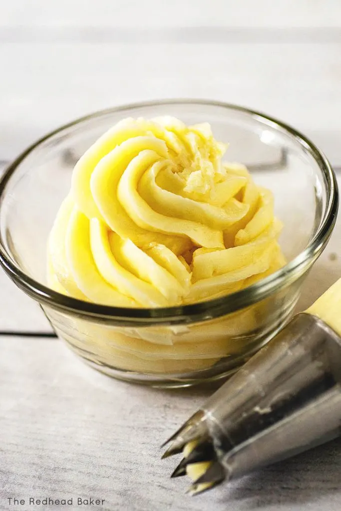 A swirl of swiss buttercream in a clear glass dish next to a piping bag fitted with a star tip
