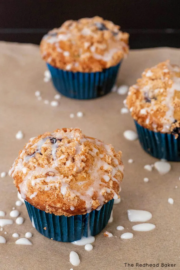 Three blueberry muffins with crumb topping and glaze on parchment paper