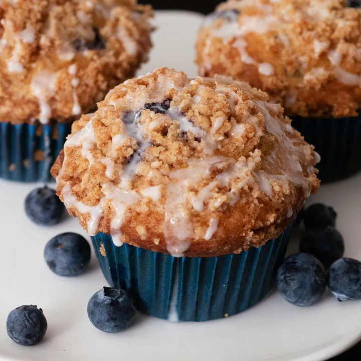 Three blueberry buttermilk muffins on a white serving stand alongside fresh blueberries
