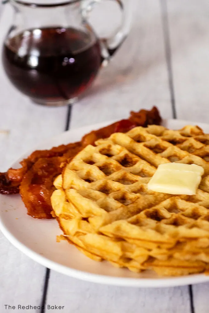 A plate of waffles topped with butter beside a few strips of bacon