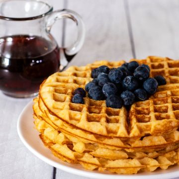 A stack of buttermilk waffles topped with blueberries in front of a jar of maple syrup