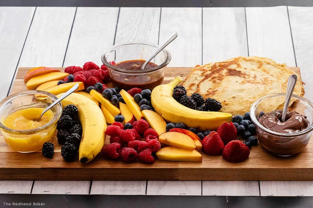 A horizontal photo of a full crepe board with crepes, three sauces and lots of fruit