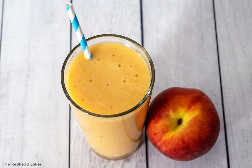 A glass of peach smoothie next to a fresh peach