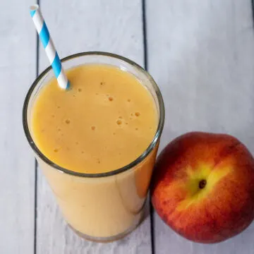 A glass of peach smoothie next to a fresh peach