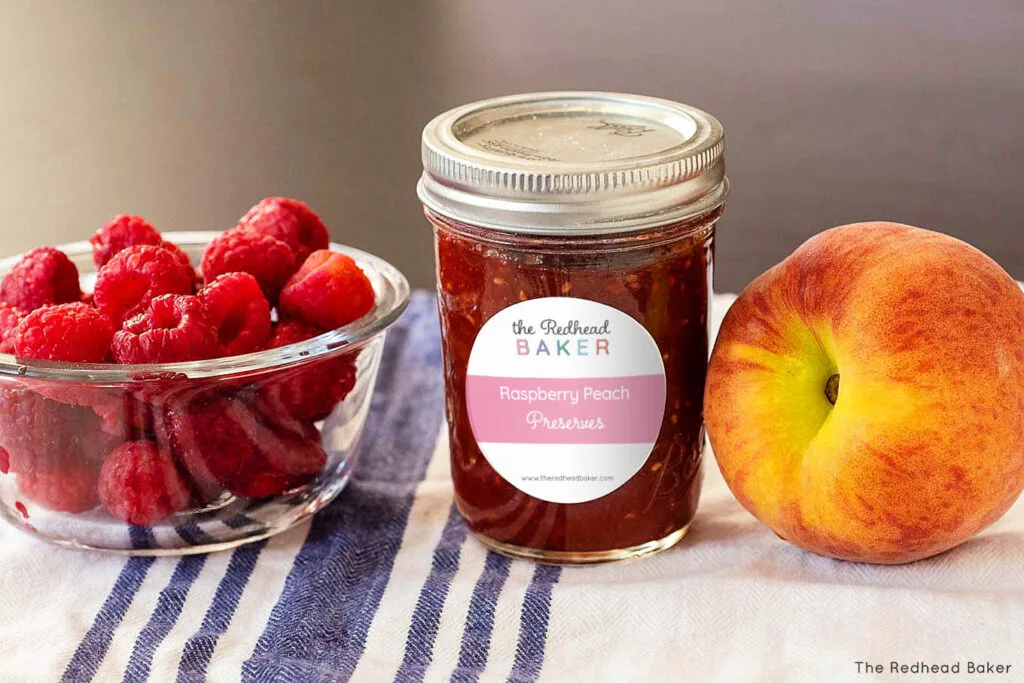 A can of raspberry peach jam in between a bowl of fresh raspberries and a fresh peach