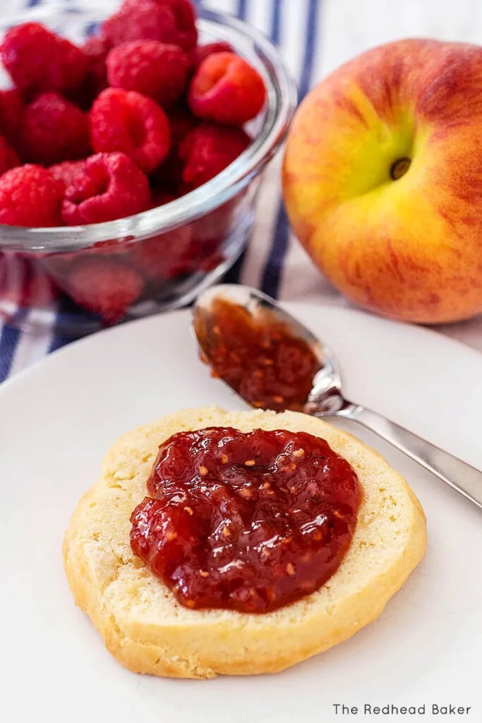 Half a shortcake spread with jam in front of a spoonful of jam, a bowl of raspberries and a fresh peach